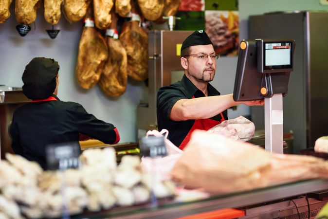 Man in butcher shop serving a customer at register with colleague behind the counter