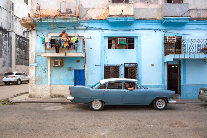 Side view of man riding a classic blue car beside blue building in Havana, Cuba
