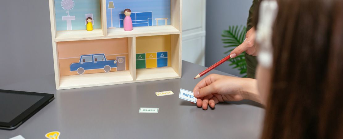 Unrecognizable teacher teaching recycling with a toy house to a female student in an ecology classroom