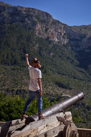 Male traveler standing on ancient cannon and showing triumph gesture