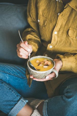 Woman in yellow shirt and jeans eating from cup of squash soup