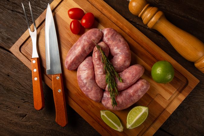 Top view of raw sausages arranged on wooden board with rosemary, lime and tomatoes