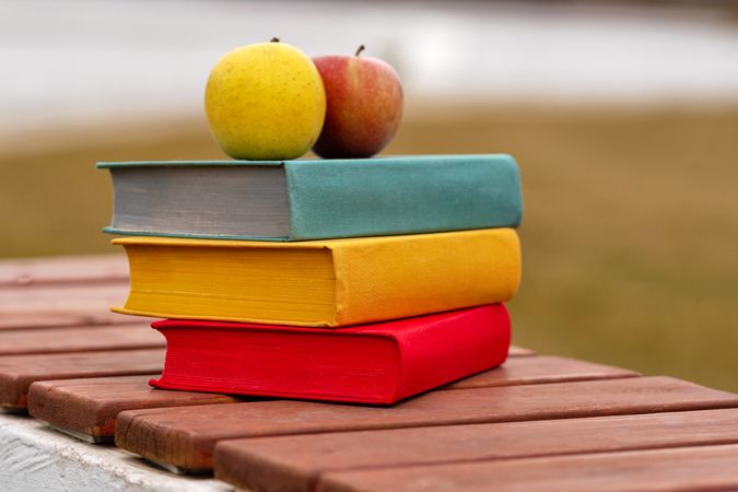 Colorful books stacked outdoors with apples placed on top.