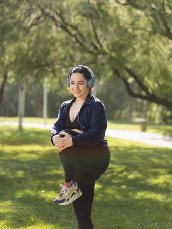 A young woman performs exercises and stretches of legs, biceps and buttocks in the morning in the park, while listening to music