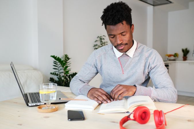 Black student reading book at table with laptop