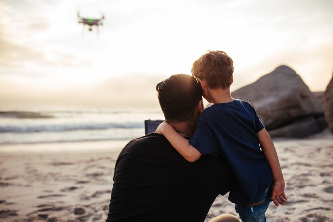 Young child hugging his dad while looking at drone over beach