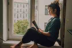 Interested woman reading book at window sill at home
