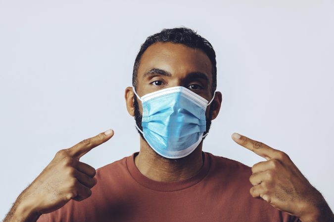Male looking at camera while pointing at face while wearing medical face mask in studio shoot
