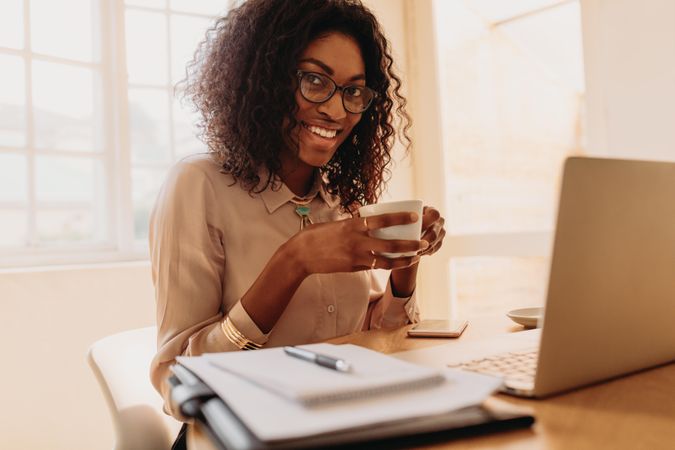 Smiling woman entrepreneur sitting at home enjoying coffee while working on laptop