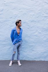 Asian male standing in front of wall in blue shirt and jeans, vertical