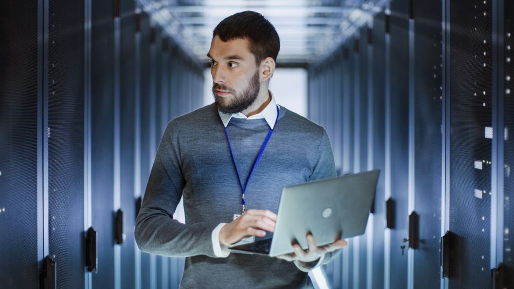 Male Server Engineer Works on a Laptop in Large Data Center.