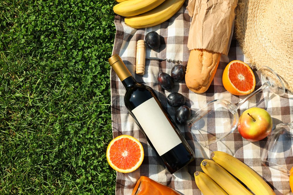 Bottle of wine, baguette, fruits, straw hat and towel on grass, space for text