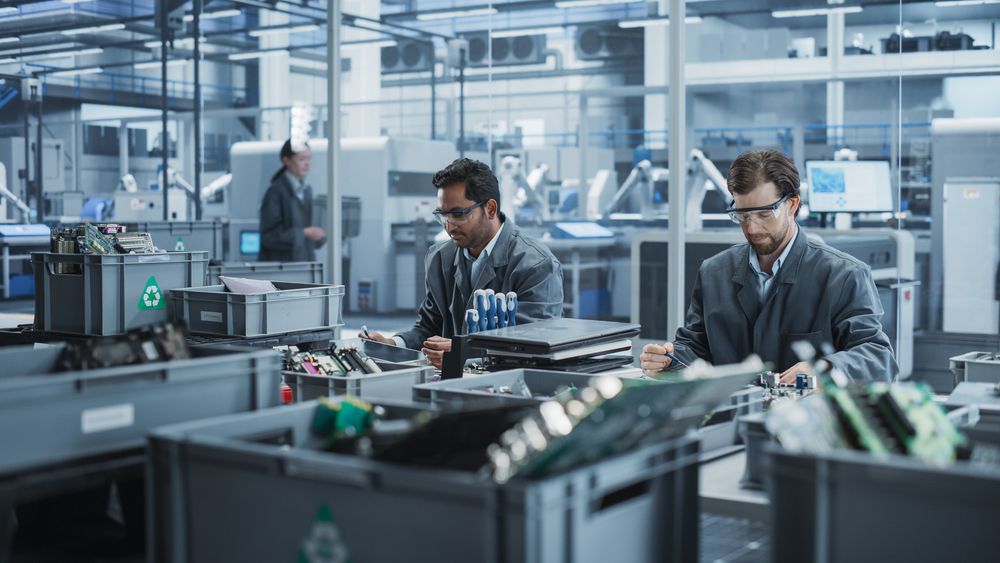 Indian And White Male Workers Taking Apart Laptops To Recycle For Production Of High-Tech Devices At Automated Electronics Factory With Robotic Arms.