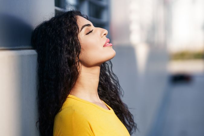 Profile of female with curly hair leaning back on grey wall outside, copy space