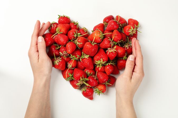 Female hands touch heart made of tasty strawberry on plain background, top view. Summer berry
