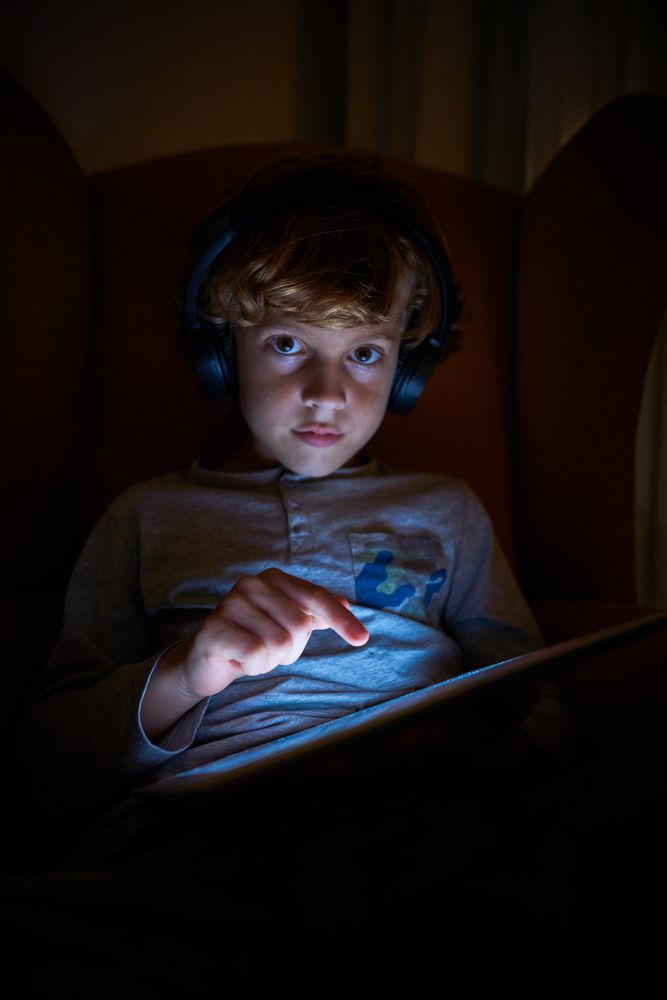 Vertical photo of a child sitting with helmets on a sofa touching the screen of an ipad and looking frontally at night
