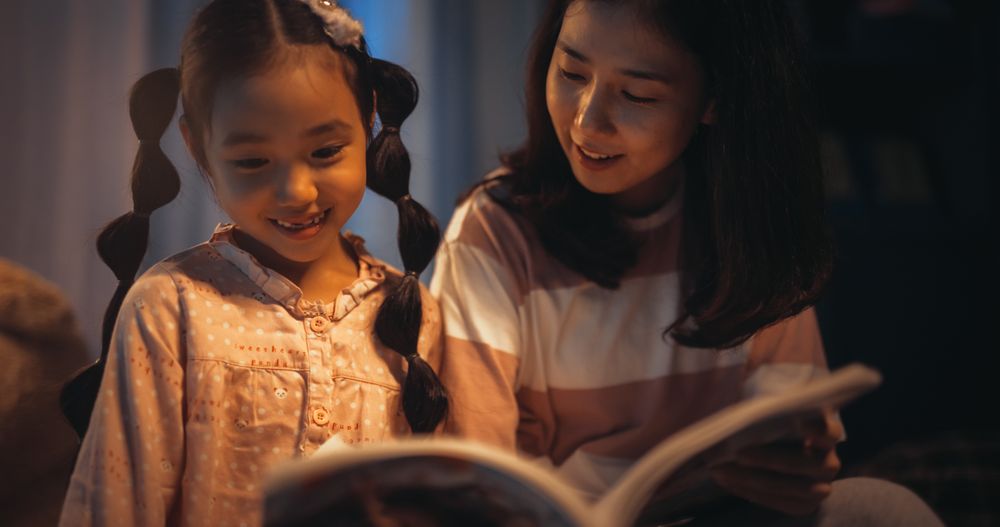 Korean Mother Reading a Fairytale to Her Lovely Small Daughter in Bed Before Going to Sleep.