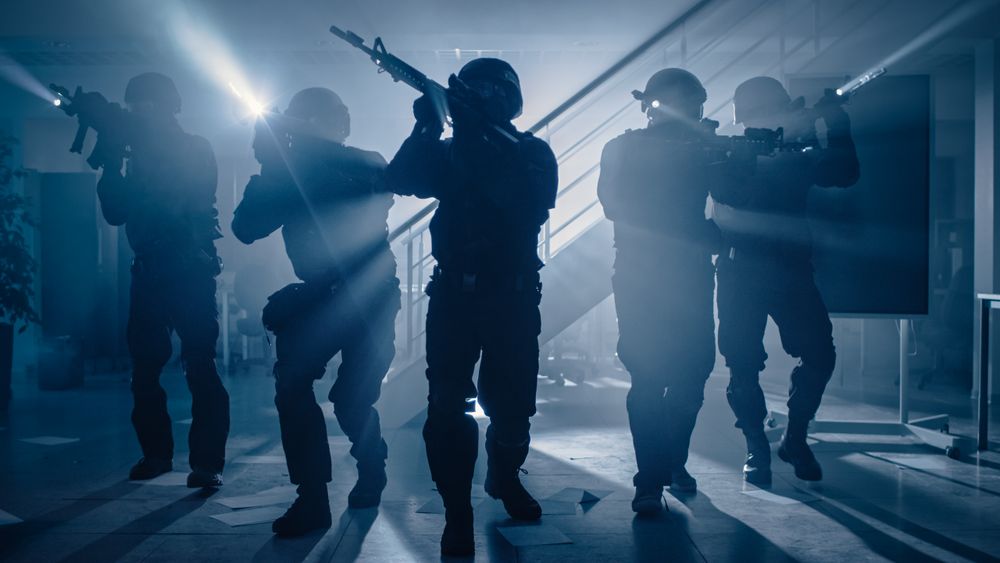 Masked Team of Armed SWAT Police Officers Move in a Hall of a Dark Seized Office Building with Desks and Computers.