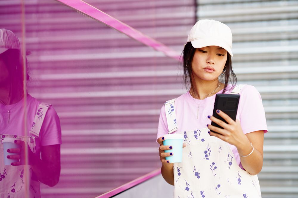 Trendy Chinese woman walking outside in coveralls holding to go coffee and smart phone