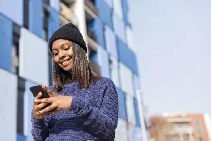 Female in hat and blue sweater checking cell phone outside blue building