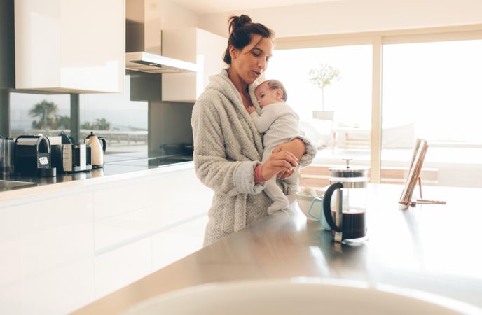 Woman in bathrobe holding her son and making baby food in kitchen