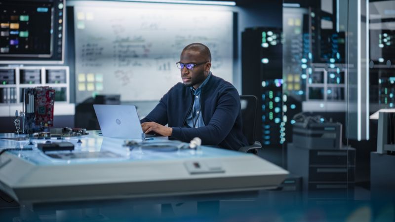 Portrait of a Male Specialist Using Laptop Computer to Improve Electronic Circuit Board in Modern Office. Black Engineer Thinking and Typing Ideas for Artificial Intelligence Project