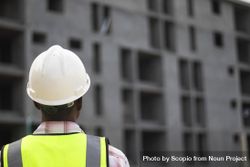 Back view of man wearing vest and bump cap standing beside a building under construction 0ypKjb