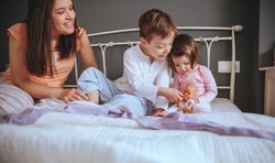 Portrait of happy children feeding doll with a cookie sitting in bed with their mother