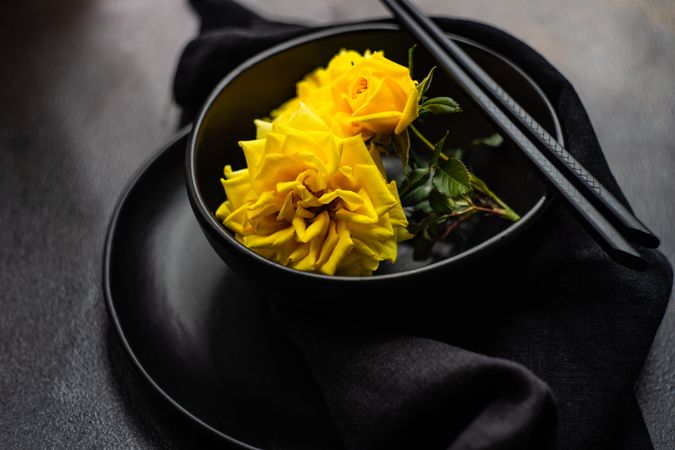 Dark table setting with yellow roses and chopsticks over bowl