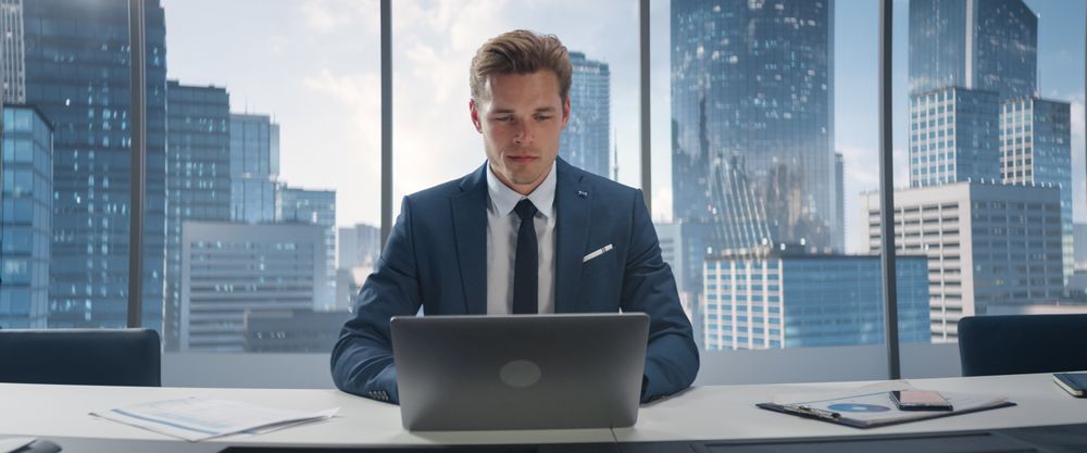 Portrait of Successful White Businessman Working on Laptop Computer in His Big City Corner Office.