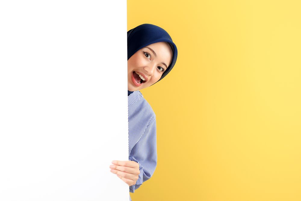 Happy Smiling Young Asian Muslim Woman with Hijab Peeking Out Behind a Wall