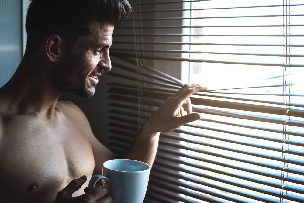 Man standing by a windows blind with a cup of coffee