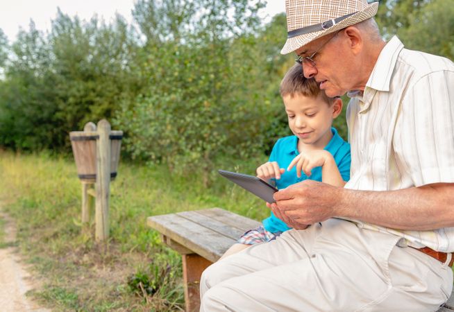 Grandchild and grandfather using a tablet sitting on bench