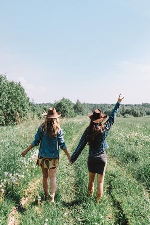 Two women wearing hats and holding hands and walking on grass field
