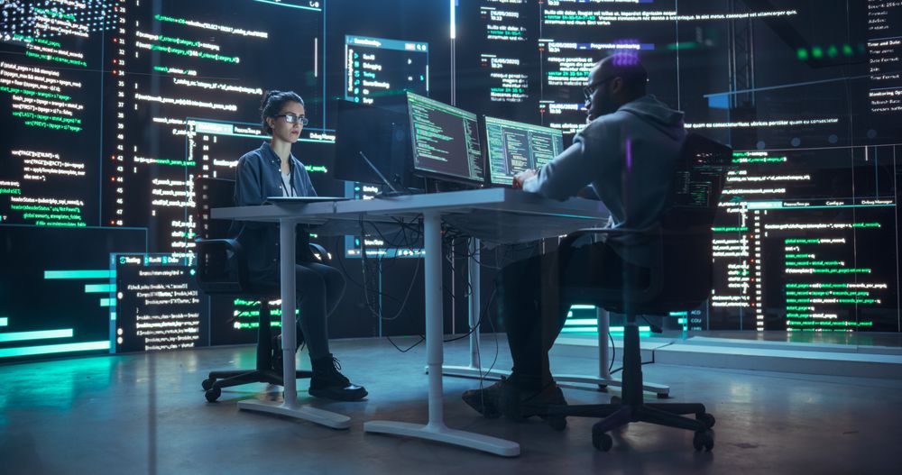Diverse Young Man and Woman Working on Desktop Computers, Surrounded by Big Screens Displaying Code Lines.