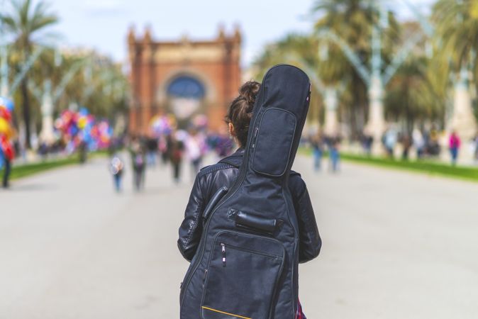 Rear view of a young hipster woman walking in a park in sunny day while carrying a guitar on back