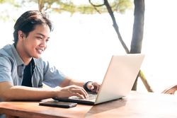 Happy Asian Young businessman work at outdoor with laptop. Concept of remote and freelance work. Young successful man wearing casual blue cloth.