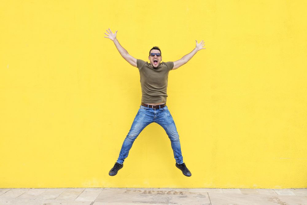 Excited male standing outside jumping in front of yellow wall with open arms