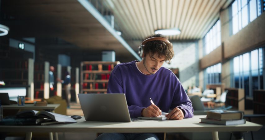 White Student Writing an Essay on Laptop Computer, Thinking About the Topic, Brainstorming, Finding Solutions. Young Stylish Man Studying in a Public Library, Listening to Lecture in Headphones