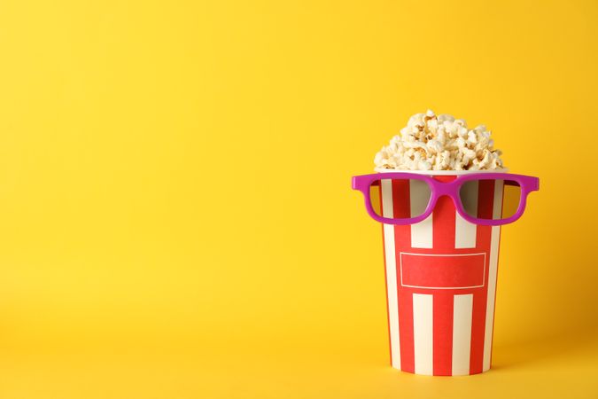 Striped bucket with popcorn and 3d glasses on yellow background, copy space