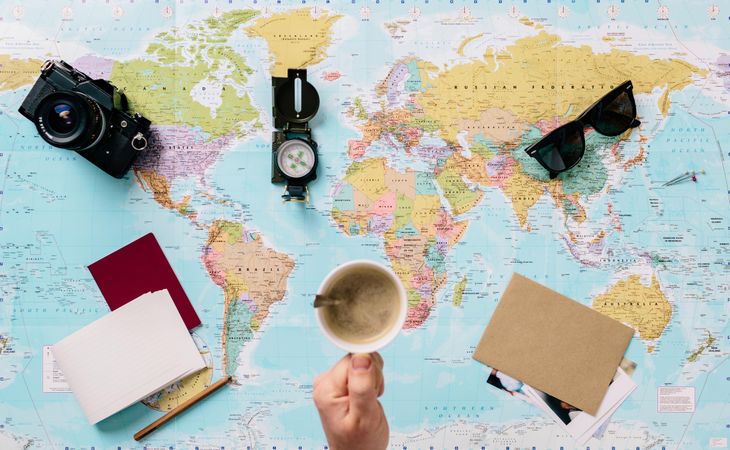 Person holding a cup of coffee over the world map with camera and compass over it