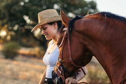 Young woman in hat walking with her horse in the countryside at sunset