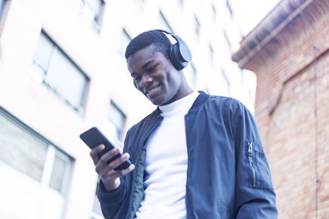 Smiling Black man standing outside, wearing headphones and looking down at his phone