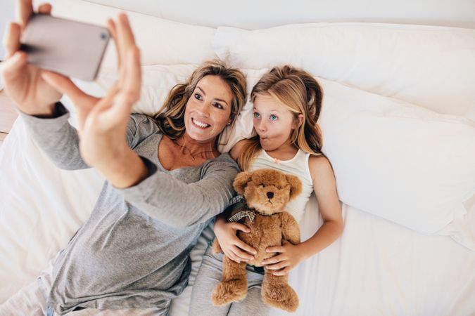 Woman taking selfie with little girl holding teddy bear in bed