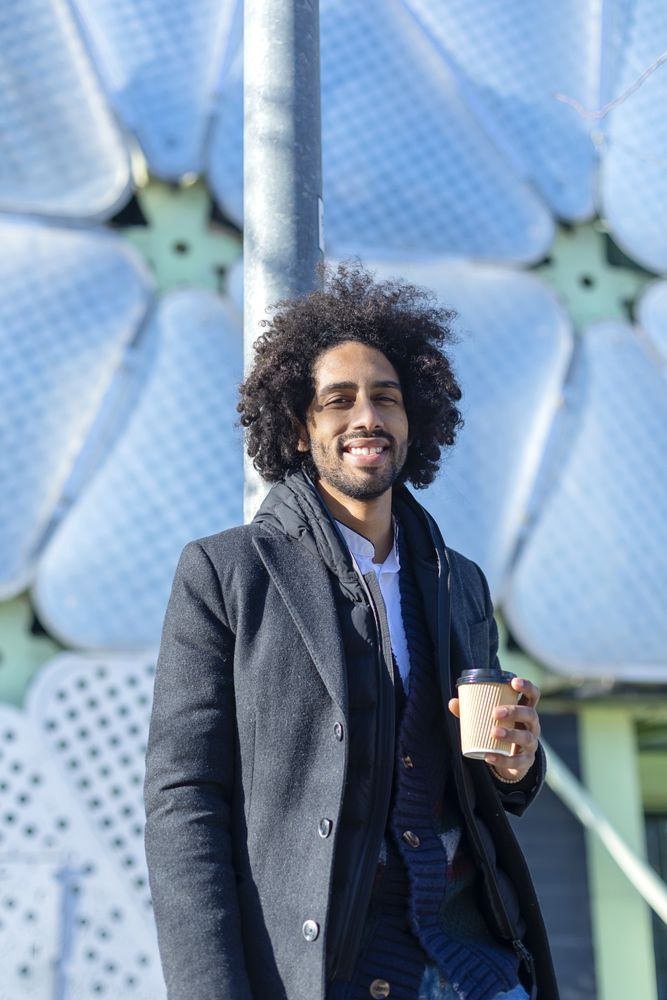 Portrait of cheerful Black man in jacket with warm beverage standing outside leaning on pole