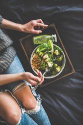 Woman in jeans sitting with wooden tray of vegetarian bowl, smoothie on side, vertical composotion