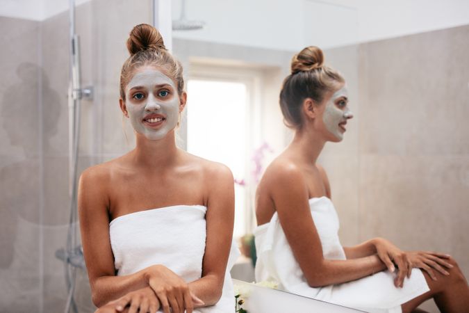 Beautiful young woman with facial mask sitting in bathroom and smiling