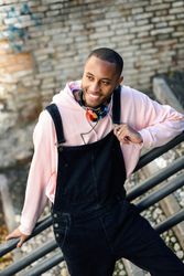Smiling male in overalls and pink hoodie leaning on railing of stairs outside