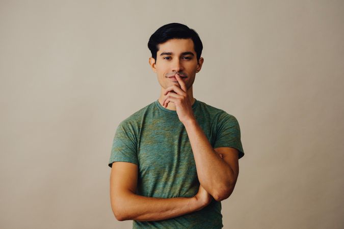 Contemplative Hispanic male standing in neutral room with arms crossed and finger over mouth