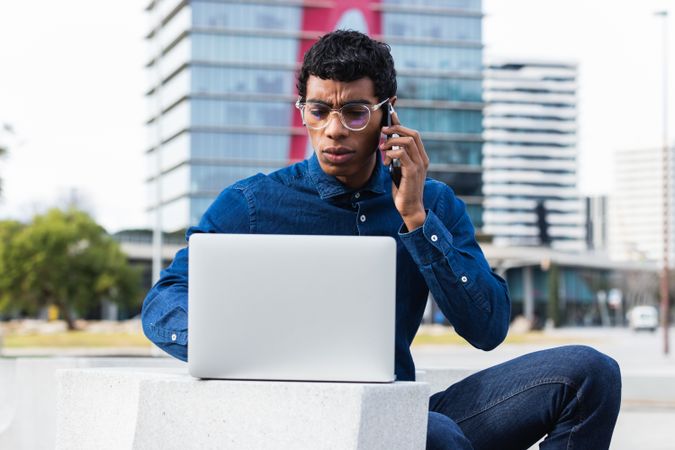 Serious hispanic businessman freelancer working on laptop and talking on mobile phone in city